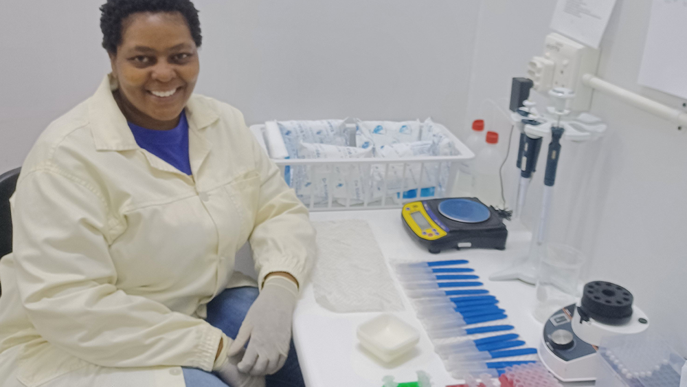 A smiling university intern named wearing a lab coat and protective gloves, sitting at a workstation organized with scientific equipment, pipettes, and tubes.