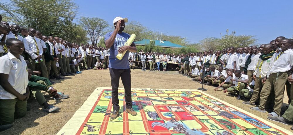 An instructor holding a rolled-up poster standing on a large educational board game mat surrounded by a crowd of school students outdoors.