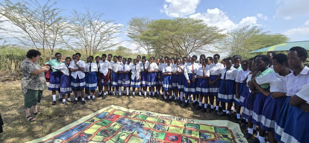A large group of school students in blue uniforms listening to an instructor outdoors, around an educational floor mat.