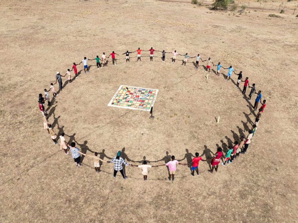 Aerial view of a large group of people holding hands in a large circle around a colorful mat in an open field.