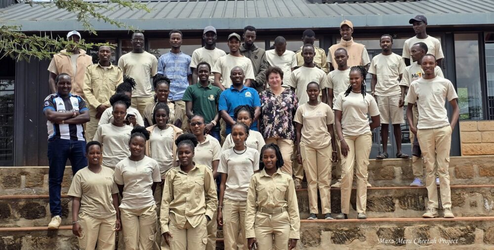 A large group photo of the Mara-Meru Cheetah Project team standing on stone steps outdoors in front of a building, wearing matching field uniforms.